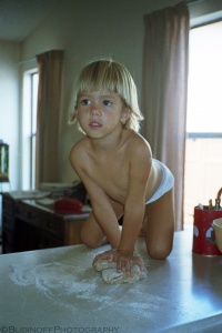 A portrait photograph of a young child kneeling on a kitchen countertop applying pressure with her hands to knead bread dough. The girl glances to the photographer's left and appears engaged in her task. Flour is spread around the dough ball and kitchen windows and drapery are defocused in the background. Budinoff Photography