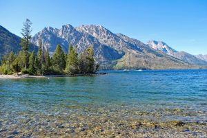 This Jenny Lake Grand Tetons perspective photo in landscape format captures the view across a lake from the shore with pine trees and racing ferry boats in the middle ground and rugged mountains in the background. The rocky bottom of the lake can be seen through the clear shallow water in the foreground. Blue skies with a few streaks of faint white clouds silhouette the mountain peaks.
