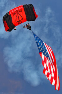 A color portrait oriented photo taken almost directly beneath a parachutist descending towards the ground. The ram air parachute is colored with broad black and red stripes. A spade shaped symbol with the words Airborne above it are visible on the middle red stripe. The parachutist is suspended below the parachute by several ropes. The parachutist is dressed in black clothing but his bare arms are visible. He wears a black helmet with a camera mounted on the top. Below the parachutist is a large USA flag. The background is comprised of blue sky with some white smoke visible. Budinoff Photography