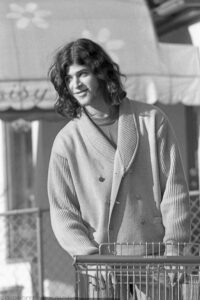 A grayscale film photo taken in 1972 of the upper two thirds of a young man standing in front of a traditional wire grocery store shopping cart. The man has long dark hair, a slight beard, and wears a light shaded double breasted long sleeve cardigan sweater with a dark shirt underneath. The man is facing the camera, smiling and gazing to his right as his hands hold the front of the cart. The store name, Grand Union, is written on the handle. The background is not focused but reveals a scalloped edge sun awning over a storefront and a low chain link fence. Budinoff Photography