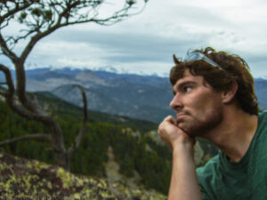 A color landscape oriented photo taken outdoors while hiking in Chautauqua Park in Boulder, Colorado. There is no focus point in the image, all details are slightly defocused. Dominating the photo is the profile of a man's face and forearm as he gazes to the left of the frame. The man has wavy dark brown hair and his clear plastic glasses rest on the top of his head. The man has a short beard and his chin is resting on the palm of his left hand. He wears a short sleeve dark green t-shirt. In the foreground are light green lichens which create a splotchy pattern on a dark brown rock. In the middle ground, dark green conifer trees cover a ridge. The curvy branches of a sparsely foliated conifer are in the middle ground on the left side of the photo. In the far ground are more conifer covered hills with some tan rock outcrops visible. On the horizon are the snow covered peaks of the Rocky Mountain's front range. Above the horizon are clouds that are white, gray, and dark gray. They do not look like they will cause rain. Budinoff Photography