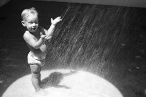 A landscape oriented black and white image of a toddler standing in a shower room with direct sunlight illuminating him and the shower spray. The bottom of the frame is comprised of a half circle of bright sunlight that is streaming through an opening in the ceiling. The young boy is standing facing right and is illuminated by the sunlight with his feet and shadow inside the half circle of light. The boy wears a bathing suit over his diaper. He holds his palms up and in front of him letting the shower spray hit his hands and his stomach. He faces the camera with a half open mouth and is squinting because of the bright sun and the shower spray. The shower spray emanates from the top right of the frame and travels towards the lower left of the frame. The shower head is not visible. The shadowed cement floor is blotchy and dark. A sliver of shower wall is brighter than the floor and is visible in the upper right of the frame. Budinoff Photography