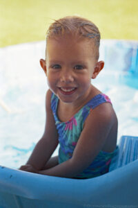 A portrait oriented photo of a young smiling girl looking at the photographer while sitting on a step in a small kiddie pool. The girls head occupies the upper third of the frame, her shoulders in the middle of the frame and her arms the lower part of the frame. The girl is in the shadows but the sun brightly illuminates the defocused pool and water behind her. She wears a blue patterned swim suit and has wet slicked back light colored hair. The blue rounded edge of the kiddie pool is at the bottom of the frame. Above the girl is defocused dormant grass that appears tannish yellow. Budinoff Photography