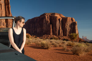 A landscape oriented color photograph of a fair skinned woman standing by an open truck door with Elephant Butte in the background. The woman and open white trimmed car door are to the left of the frame. The woman wears a black tank top and sunglasses, her hair is clipped up in back. The vibrant red rocks of prominent Monument Valley formations fill the right two thirds of the frame. A clear blue early morning sky with a few stratified clouds fills the top and middle right of the frame.