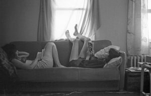 A landscape oriented black and white image of two woman reading on a couch in a room only illuminated by light coming through a window behind them. Both woman are reclined with pillows against the arms of the couch. Both are wearing shorts. The woman on the right has her legs up over the top of the couch and her bare feet are silhouetted by the bright light shining through the sheer curtain covered window. She is reading a paperback book titled The Godfather. The woman on the left has her legs bent and her feet are on the cushion of the couch. The vertical pipe elements of a radiator are visible to the right of the couch. Budinoff Photography