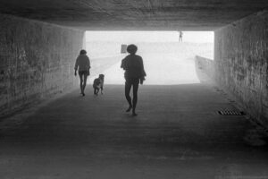 A landscape oriented grayscale photograph taken in s shady tunnel that opens to the bright sand of a beach on Long Island, NY. Two people are walking in shorts and barefoot in the tunnel, one with a dog on a leash, the other carrying a towel over his shoulder. The film exposure is grainy at the bright tunnel exit where a lone figure stands on top of a sand dune with their right arm raised. The people in the tunnel are deeply shaded so they appear almost like silhouettes. Budinoff Photography