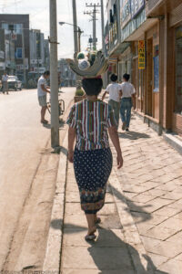 A portrait oriented color photograph of a woman's back while she is walking on a sidewalk with a bundle of folded textiles on her head. The woman is wearing a dark blue skirt with marble sized white dots and a short sleeve shirt with various colorful vertical stripes. Two boys with white short sleeve shirts and blue jeans walk closely together about 20 feet in front of the woman. Storefronts with signs written in Korean are visible on the buildings that line the right side of the frame, a fairly empty street is on the left. Gray colored metal electrical suspension posts are visible with some wires randomly reaching across the street. Budinoff Photography