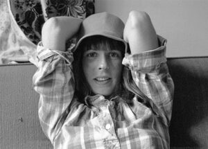 A landscape oriented grayscale photograph of a woman sitting on a couch. The woman's upper torso and head fill the frame. She wears a plaid shirt with the sleeves rolled up. Her elbows are raised to head level and her hands disappear behind her head. The woman has dark hair and her bangs just cover her eyebrows. She is smiling and her upper teeth are exposed. She looks at the camera and her eyes appear to be light colored. She wears a floppy hat made of smooth fabric. Her arms compress the brim of the hat towards her head. The couch fabric is course with vertical ribs. A long horizontal bead of upholstery marks the top back of the couch. A flower patterned fabric remanent is visible to the upper left. The light colored wall is visible above the couch back to the right. The scene is illuminated by diffused natural light from the left. Budinoff Photography