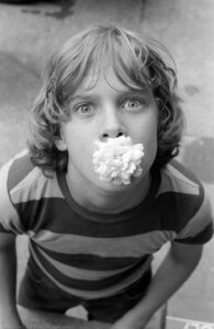A black and white close up photograph in portrait orientation of a adolescent boy taken from slightly above his head pointed down at his face while he stares into the camera. The boy's mouth is holding a light colored floribunda rose flower. He has slightly long light colored hair and bright blue eyes. He wears a broad striped short sleeve shirt and jeans. The blurred background that surrounds the boy appears to be a concrete surface. Budinoff Photography