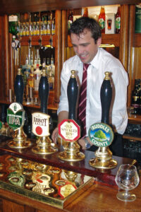 A portrait orientation photograph of a male Eagle Pub bartender smiling while pulling a draft beer behind a bar in Cambridge, UK. The bartender wears a white long sleeve collared shirt and a red striped tie. Four dark handled tall tap pulls are in front of the bartender each with it's distinctive decorative brand name labels. Distorted reflections of those name tags are visible in a shiny flat surface on the bar between the taps and the photographer. Budinoff Photography.
