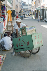 A portrait photograph of an older male wearing a white baseball cap and tee shirt leaning against a green wooden two wheeled ice cream cart. Red hand written Korean lettering appears on the front and side of the cart. A rectangular green box with clear sides and a hinged top is filled with stacked light tan ice cream cones. A man in a white long sleeve shirt squats in the street behind the ice cream man with his back to the camera. Women's clothes can be seen hanging on the left side of the photo in a store front. Other people can be seen walking in the distance on the same street. Budinoff Photography