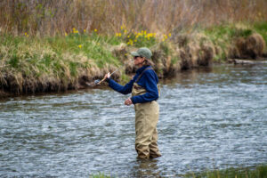 A landscape oriented color photograph of a fly fisherman standing in calf high water South Platte River water near Deckers, Colorado on a cloudy day. The woman is centered in the frame and is casting her lure towards the left of the frame. She wears tan colored waders, a blue long sleave sweatshirt and a gray baseball hat. A small portion of green grass along the near bank is on the bottom of the frame. The blue gray water around the woman is flowing towards her right. In the far ground the other side of the bank is also comprised of green grass near the water and taller brown bushes to the top of the frame. The foreground and back ground are slightly defocused. Budinoff Photography