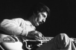A close-up black and white photograph of jazz guitarist Pat Martino while he was playing at Grendel's Lair nightclub in Philadelphia. The artist is sitting on a chair with his right leg crossed and his guitar is resting in his lap. His right hand is strumming the strings. The artist appears to be looking at his left hand but the hand is obscured by his left knee. The subject is illuminated by a spot light above and to the right of the frame. This causes deep shadows on the artists face from his cheek bones. The artist is wearing a mock turtleneck sweater over a light colored collared shirt. Budinoff Photography