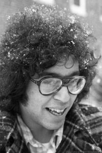 A black and white portrait of a smiling young man with thick eye glasses and long dark curly hair during a snow storm. Snow can be seen falling in the defocused background and snowflakes can be seen in his hair. He wears a plaid colored shirt covered with a plaid overcoat. A brick building can be seen in the background with partial frames of two adjacent windows apparent. Budinoff Photography