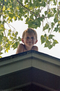 A close-up of a shirtless young boy posing at the pinnacle of a roof top. He is grinning contentedly and his shaded head is framed by green leaves on tree branches. Budinoff Photography
