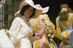 A landscape orientation color photograph of a bride, maid of honor and bride's maid taken on the stairs at the entrance to a church on a sunny morning. The bride is wearing a cream colored gown with a high collar. She has turned smiling towards the photographer and holds her train in her right hand and a white rose bouquet in her left. She is wearing a floppy broad brimmed hat that has lattice openings on the brim. Her wedding party have turned to enter the church. They wear yellow and pink dresses. Budinoff Photography