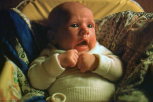 A landscape photograph taken in ambient light of a three month male wearing a white tight knit sweater. His gaze is to his left at his mother. He is cradled in a homemade quilt and leans back against his infant carrier. His face is shadowed on his right side but his ear is still visible. Budinoff Photography